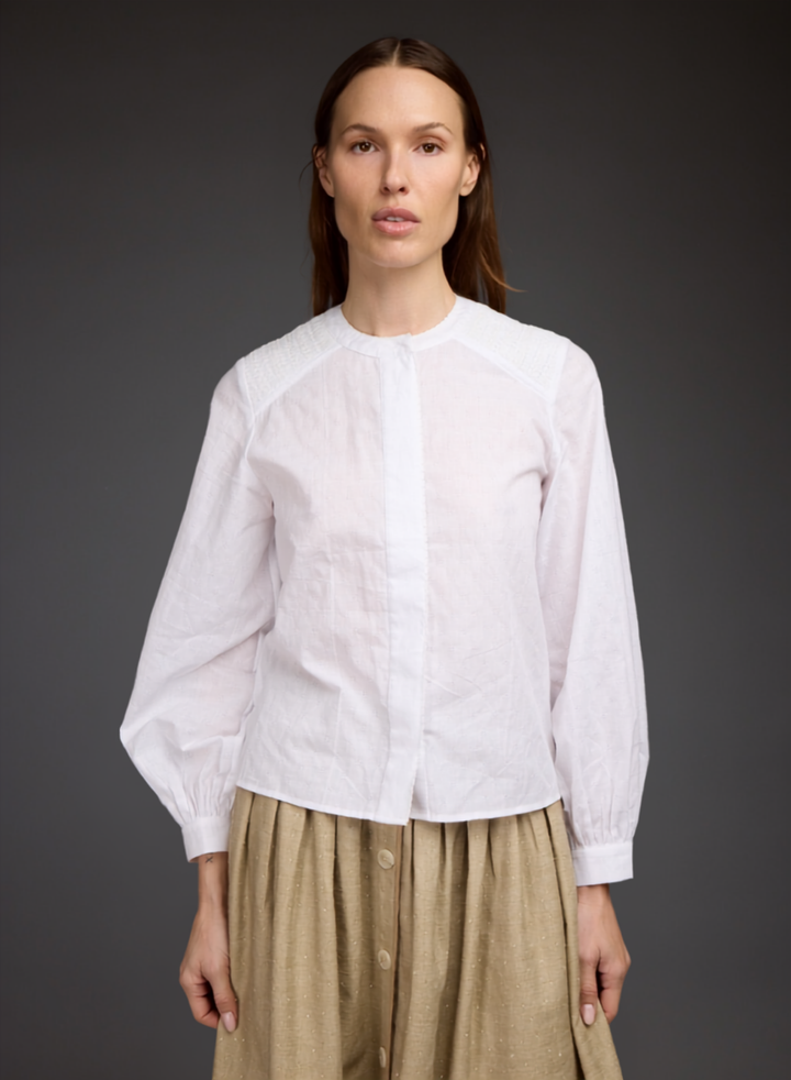 A woman modeling a white long-sleeve blouse with a round neckline and gathered sleeves, paired with a beige skirt, against a dark gray studio background.