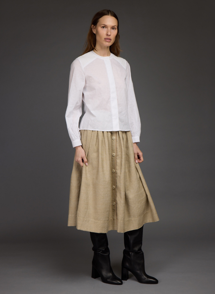 A model stands against a dark gray studio background wearing a white long-sleeve blouse with a sheer yoke and a beige button-front midi skirt, paired with black knee-high boots.
