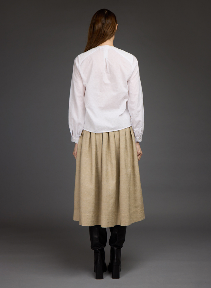 A woman shown from the back wearing a white long-sleeve blouse and a beige pleated midi skirt, paired with black ankle boots, against a dark gray studio background.