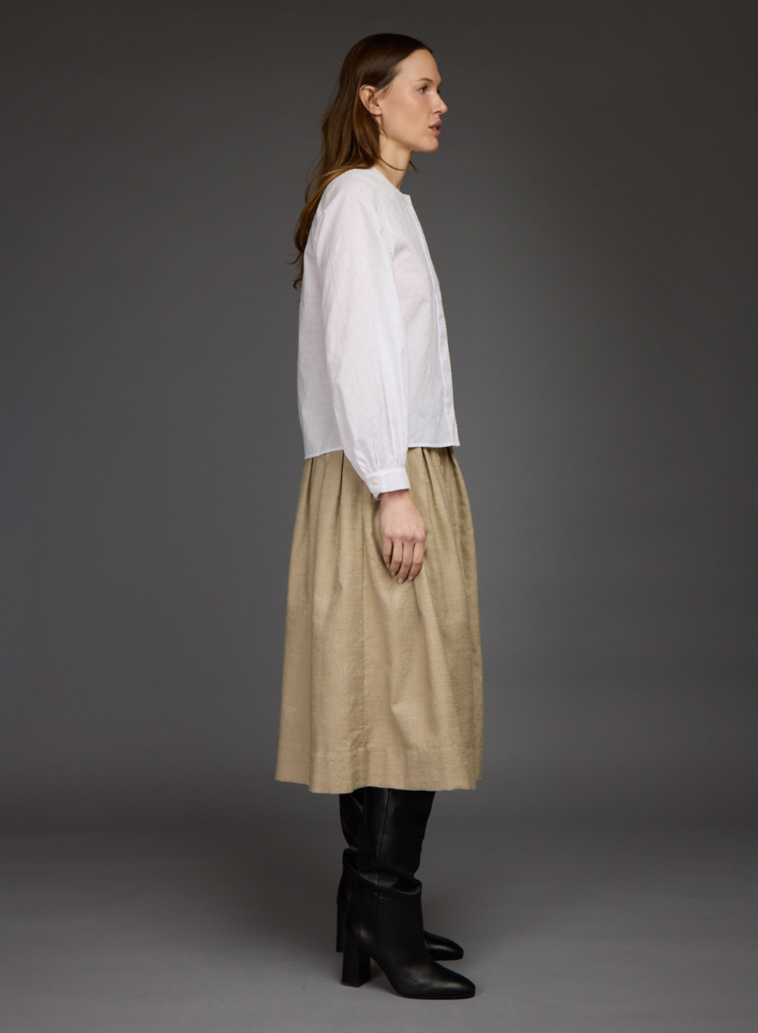 A woman stands in profile wearing a white long-sleeve blouse, a beige midi skirt, and black knee-high boots against a dark gray studio background.