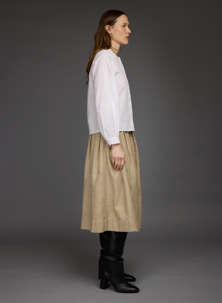 A woman stands in profile wearing a white long-sleeve blouse, a beige midi skirt, and black knee-high boots against a dark gray studio background.