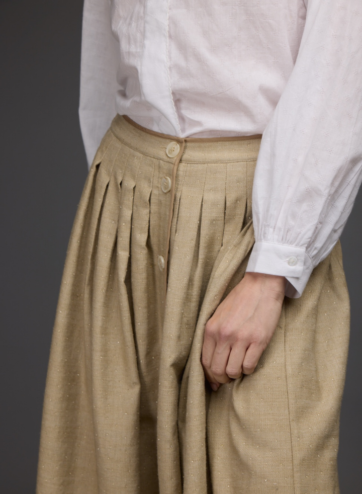 A close-up of a person wearing a beige pleated skirt with front buttons and a white long-sleeve blouse, shown against a dark gray studio background.