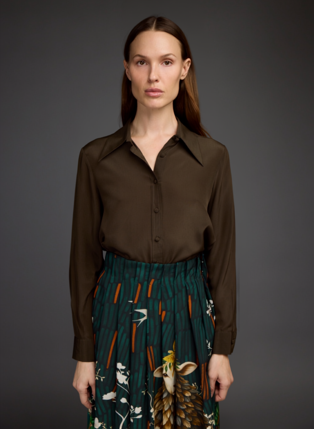 A woman modeling a dark brown long-sleeve button-up shirt paired with a dark green pleated skirt featuring a detailed bird and floral print, set against a solid dark gray studio background.