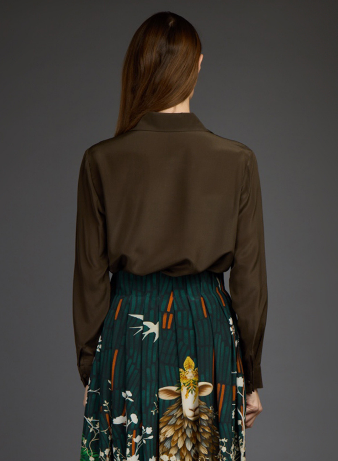 A model shown from the back wearing a dark brown satin long-sleeve blouse and a pleated dark green skirt with a detailed sheep and bird print by Love Binetti, set against a solid dark gray studio background.