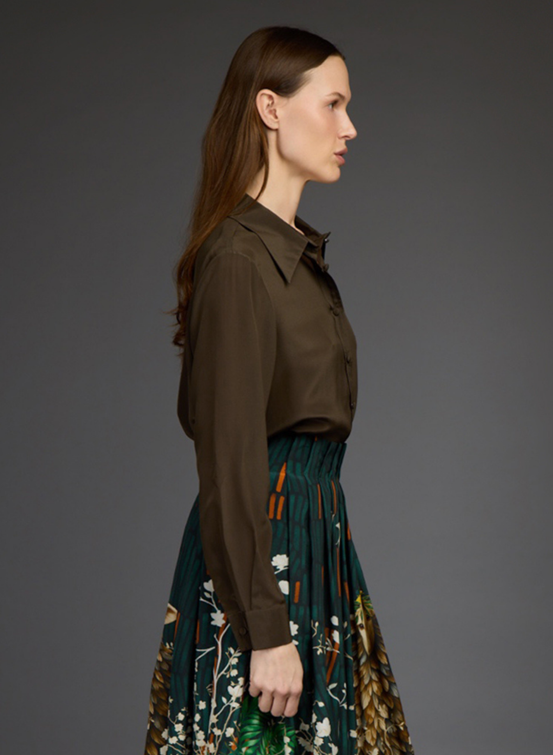 A woman shown in side profile wearing a dark brown long-sleeve collared shirt tucked into a pleated dark green skirt with a botanical print and orange accents by Love Binetti, against a solid dark gray studio background.