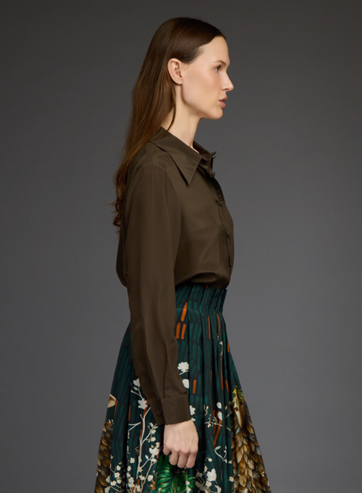 A woman shown in side profile wearing a dark brown long-sleeve collared shirt tucked into a pleated dark green skirt with a botanical print and orange accents by Love Binetti, against a solid dark gray studio background.