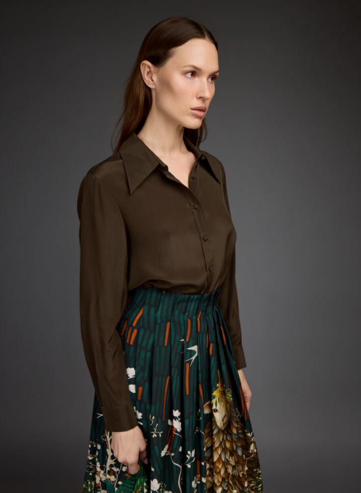 A woman modeling a dark brown long-sleeve button-up shirt paired with a dark green pleated skirt featuring a colorful floral and bird print, set against a dark gray studio background.